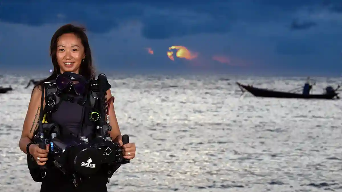 Diver holding an underwater video camera housing at sunset with a longtail boat in the background, Koh Tao, Thailand